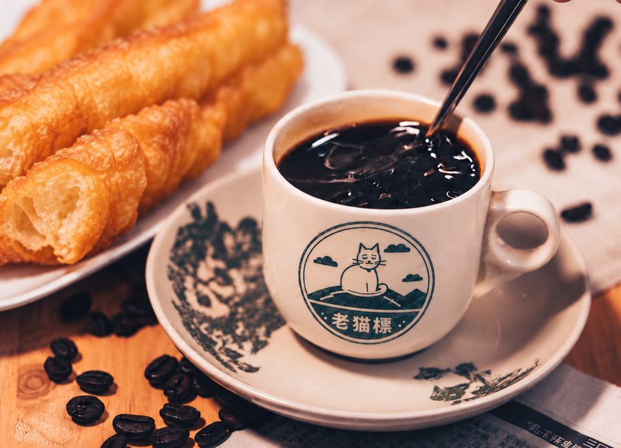 A cup of black Hainan kopi in a Lao Mao cup, with youtiao and roasted coffee beans